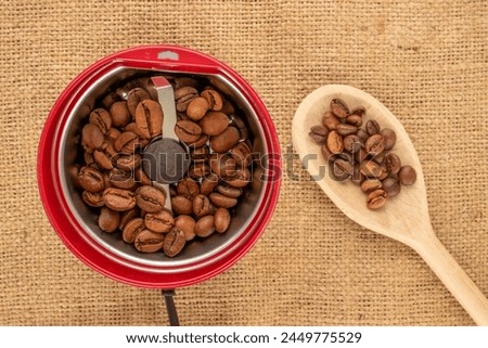 One electric coffee grinder with coffee beans and wooden spoon on jute cloth, macro, top view. Royalty-Free Stock Photo #2449775529