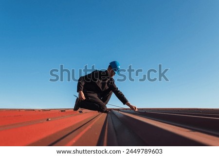 Young man worker in helmet holding meter tape on roof with blue sky. Roofer work background Royalty-Free Stock Photo #2449789603