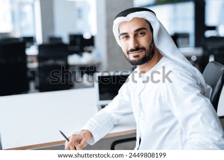 Handsome man with dish dasha working in his business office of Dubai. Portraits of a successful businessman in traditional emirates white dress turning back and smile in camera. Royalty-Free Stock Photo #2449808199