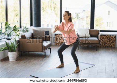 Caucasian slim young woman in sporty clothes doing squats on fitness mat at home. Fit female athlete training, stretching her legs in the living room indoors Royalty-Free Stock Photo #2449846893