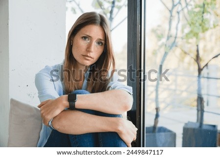 Depressed young woman thinking about breakup sitting by the window alone at home. Caucasian sad stressed girl suffering from period crumps, trauma, loneliness indoors Royalty-Free Stock Photo #2449847117