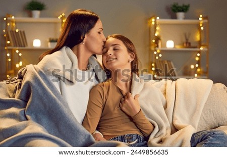 Smiling happy young mother with child girl sitting on sofa at home wrapped in cozy blanket and hugging. Joyful woman kissing her daughter indoors. Mothers day, love and care concept.