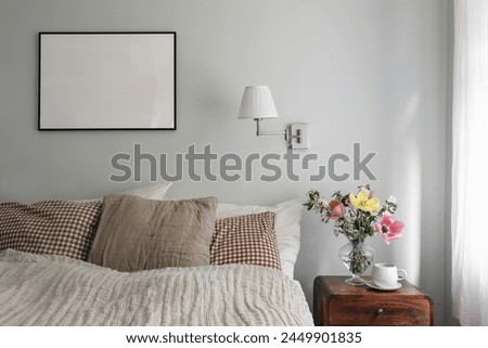 Elegant bedroom. Tulips, cherry tree blossoms bouquet in glass vase. Wooden night stand. Cup of coffee. Blank black picture frame mockup. Lamp, pleated shade on mint wall, checkered pillows.