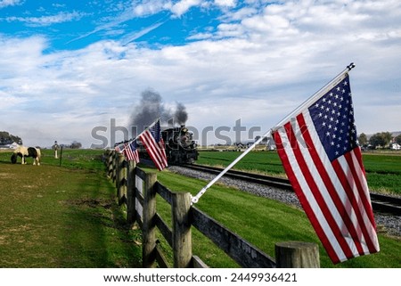 A vintage steam train chugs along the countryside, flanked by American flags on a wooden fence, evoking a sense of nostalgic Americana and historic travel. Royalty-Free Stock Photo #2449936421