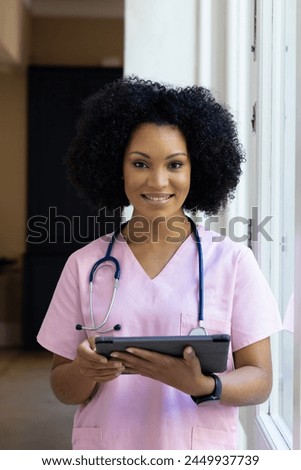 Biracial nurse holding tablet, wearing pink scrubs, standing by window at home. She has curly black hair, light brown skin, and is smiling warmly, unaltered. Royalty-Free Stock Photo #2449937739
