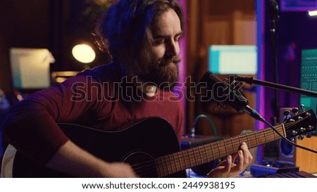 Artist with condenser microphone composing a song on guitar, working on mixing and mastering music. Producer recording tunes and adding sound effects in post production. Camera A.