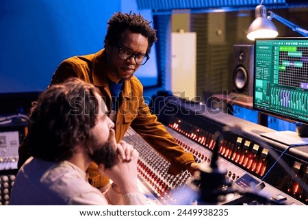 African american artist works with producer in professional studio, talking about adding new sound effects on tracks recorded. Young musician and technician editing songs with control panel desk.