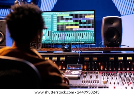 African american tracking engineer working with editing software on pc, producing new music for his album. Sound designer adjusting recordings setting and volume levels at control room panel board.