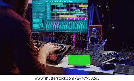 Musical performer creating mixed sounds and tunes with mixing console, greenscreen running on phone display. Artist recording new notes using equalizer and daw software on computer. Camera B.