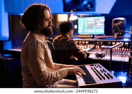 Young artist songwriter creating a new song with piano keys in studio, collaborating with audio engineer in control room. Musician singer composing music with midi controller electronic keyboard.