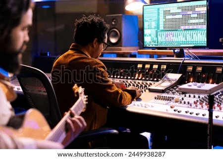Skilled songwriter artist recording a song with electro acoustic guitar tunes, making music for his pop rock album. Musician singing tracks on instrument in control room, works with audio expert.