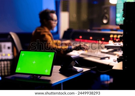 African american sound engineer editing music next to greenscreen display on gadget, operating on control room mixing console. Young producer pressing knobs and sliders in studio.