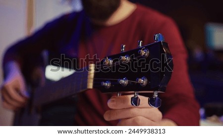 Musical performer tuning his guitar by twisting the knobs, preparing to play acoustic instrument in home studio. Artist recording sounds to create new track, using equalizer mixing gear. Camera B.