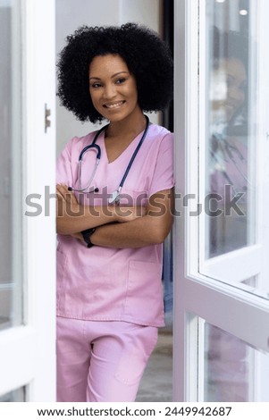 Biracial nurse standing by door at home, arms crossed, wearing pink scrubs. She has curly black hair, warm smile, and light brown skin, unaltered.