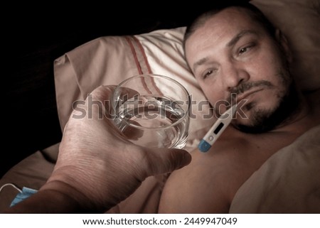 The young man feels unwell and lies in bed. He takes antipyretic pills to bring down his temperature. He regularly measures his body temperature to monitor his illness.