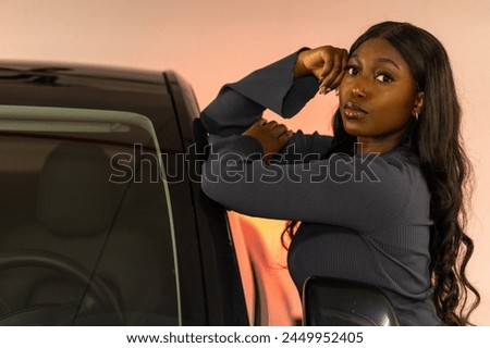 A poised African American woman leans against her car, her hand elegantly resting on the roof as if she is preparing for a drive into the sunset. The warm hues enveloping the scene suggest the end of Royalty-Free Stock Photo #2449952405