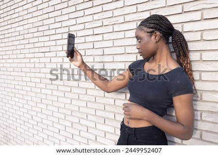 In this image, a young African woman is positioned in profile, holding up her smartphone to take a selfie. She is set against a backdrop of a white brick wall, creating a monochromatic background