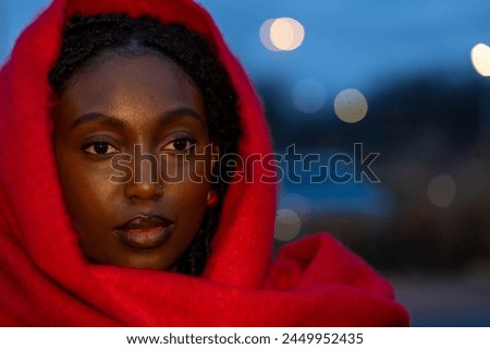 This evocative portrait features a young woman wrapped in a vibrant red scarf, her gaze penetrating and full of depth against the twilight backdrop. The scarf frames her face, drawing attention to her Royalty-Free Stock Photo #2449952435