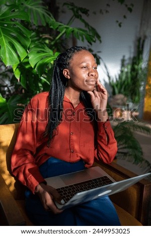 Dreaming african american distracted from work on laptop looking at window enjoy sunbeams on face sitting on chair monstera background. Relaxed black female take break from computer rest contemplative