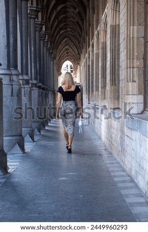 The photograph captures a solitary moment as a young Caucasian woman with blonde hair walks down a majestic gothic corridor. The symmetry of the arches draws the eye along her path, and her modern