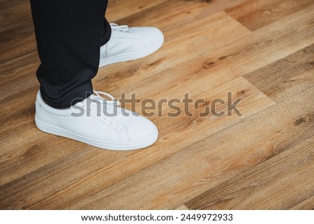 A person with white sneakers stands on a wooden floor made of brown planks. The flooring appears to be hardwood with a glossy varnish finish