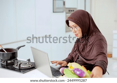 Happy smiling asian older muslim woman looking at a laptop computer in apron cooking healthy food