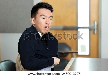 A young Asian male student in a classroom scene putting on a sweater getting dressed. The image showcases dressing for comfort in an academic setting