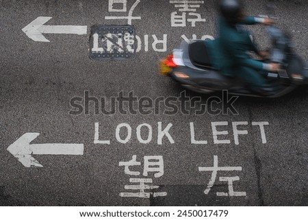 A blurred motion image of a motorcyclist passing over a 'Look Left' sign on a busy city street, capturing urban commuting and traffic safety.