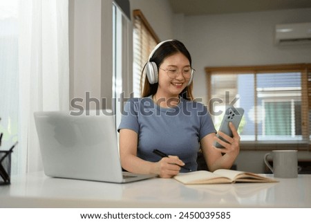 Smiling young woman typing a message on mobile phone and working with laptop at home