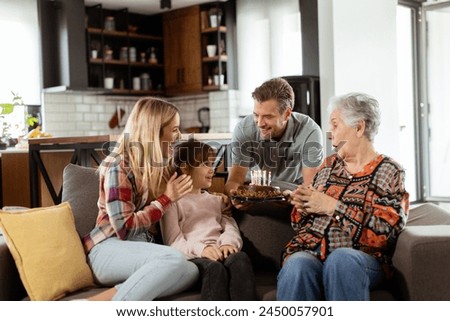 Heartwarming scene unfolds as a multi-generational family gathers on a couch to present a birthday cake to a delighted grandmother, creating memories to cherish Royalty-Free Stock Photo #2450057901