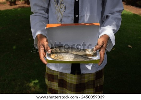 Asian Muslim adult male hand carrying a box containing soft-boned milkfish ready for consumption, Eid gift package, copy space.