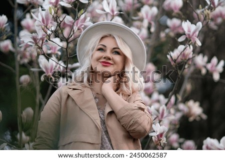 Magnolia flowers woman. A blonde woman wearing a hat stands in front of a tree with pink magnolia flowers. She is wearing a tan coat and a dress.