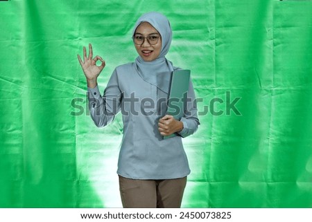 cheerful asian woman wearing hijab, glasses and blouse holding laptop and making ok gesture isolated over green background