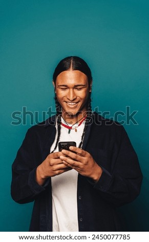 Happy man with long braided hair, wearing casual clothing and a native American necklace, standing in a studio. He smiles while reading a message on his smartphone against a blue background.