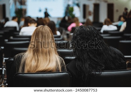 Two women are seated in a lecture room, leisurely watching a fashion presentation. The crowd is attentive as the latest trends are showcased on the screen