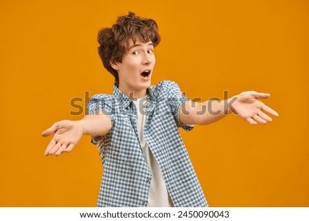 An emotional boy teenager makes his eyes wide, spreads his arms and opens his mouth in surprise. Portrait on a yellow studio background with copy space. Education. Emotions.