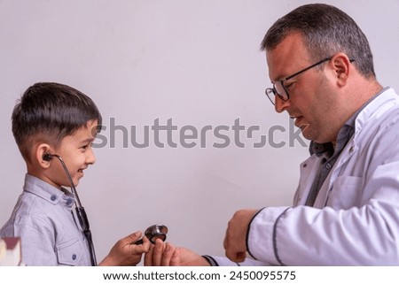 A young boy is being examined by a doctor. The boy is wearing a stethoscope and is smiling. The doctor is wearing a white coat and glasses