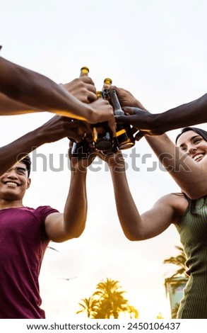 Vertical photo of happy multiracial friends having fun while chilling at sunset. Diverse young people toasting beer bottles on the beach. Celebration and summer party concept