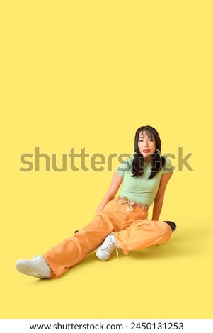 Stylish young Asian woman with bows sitting on skateboard against yellow background