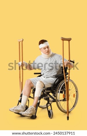 Injured young man after accident in wheelchair with crutches on yellow background