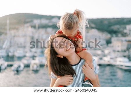 Little girl almost kisses her smiling mother on the cheek while sitting on her shoulders on the seashore