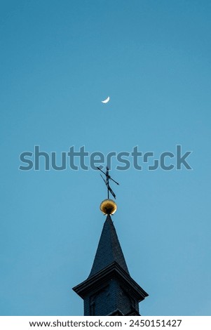 Tip of a roof with a cross of Christ and the crescent moon in the background in the daytime sky.