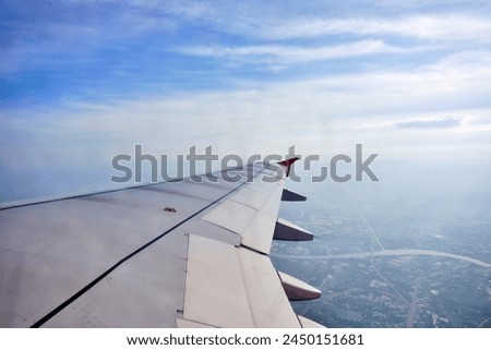a view from an airplane window that has a wing with a red winglet in the sky with clouds and a city below Royalty-Free Stock Photo #2450151681