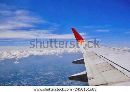 a view from an airplane window that has a wing with a red winglet in the sky with clouds and a city below Royalty-Free Stock Photo #2450151683