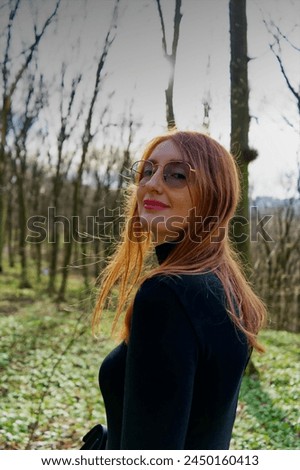 A person with obscured face stands amidst a serene forest, the sunlight filtering through bare trees creates a peaceful atmosphere, highlighting the individual’s red hair.  Royalty-Free Stock Photo #2450160413