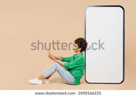 Full body young woman of African American ethnicity wear green shirt casual clothes sits near big huge blank screen mobile cell phone with area hold use smartphone isolated on plain beige background