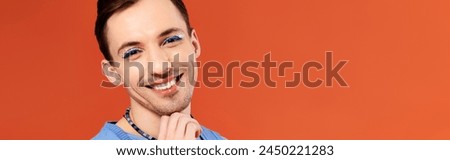 stylish joyous good looking gay man with stylish makeup posing with rainbow flag, pride month