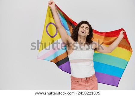 appealing joyous gay man with long dark hair posing with rainbow flag and looking at camera