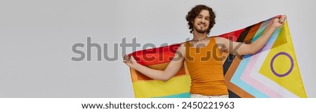 jolly alluring gay man with dark hair holding rainbow flag and smiling happily at camera, banner