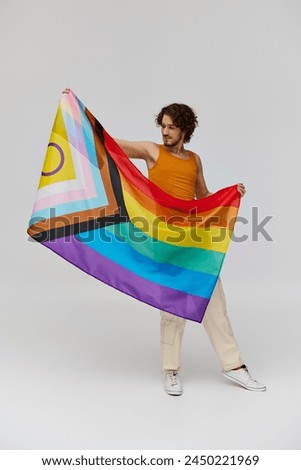 joyous young gay man with dark hair in vibrant attire posing with rainbow flag and looking away